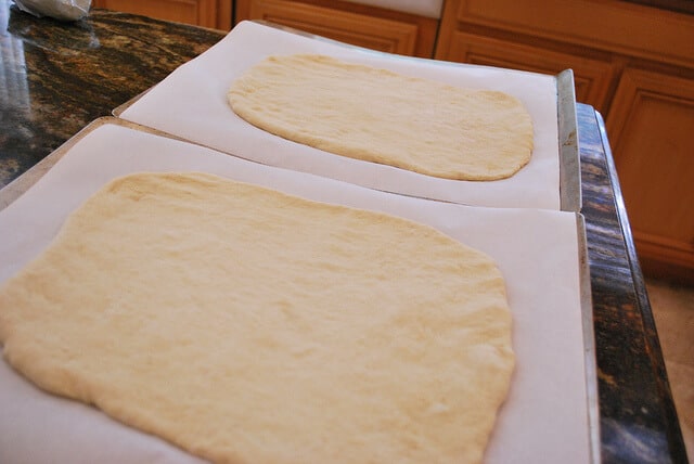 An in process image showing two pizza crusts rolled out and placed on parchment paper lined baking sheets.