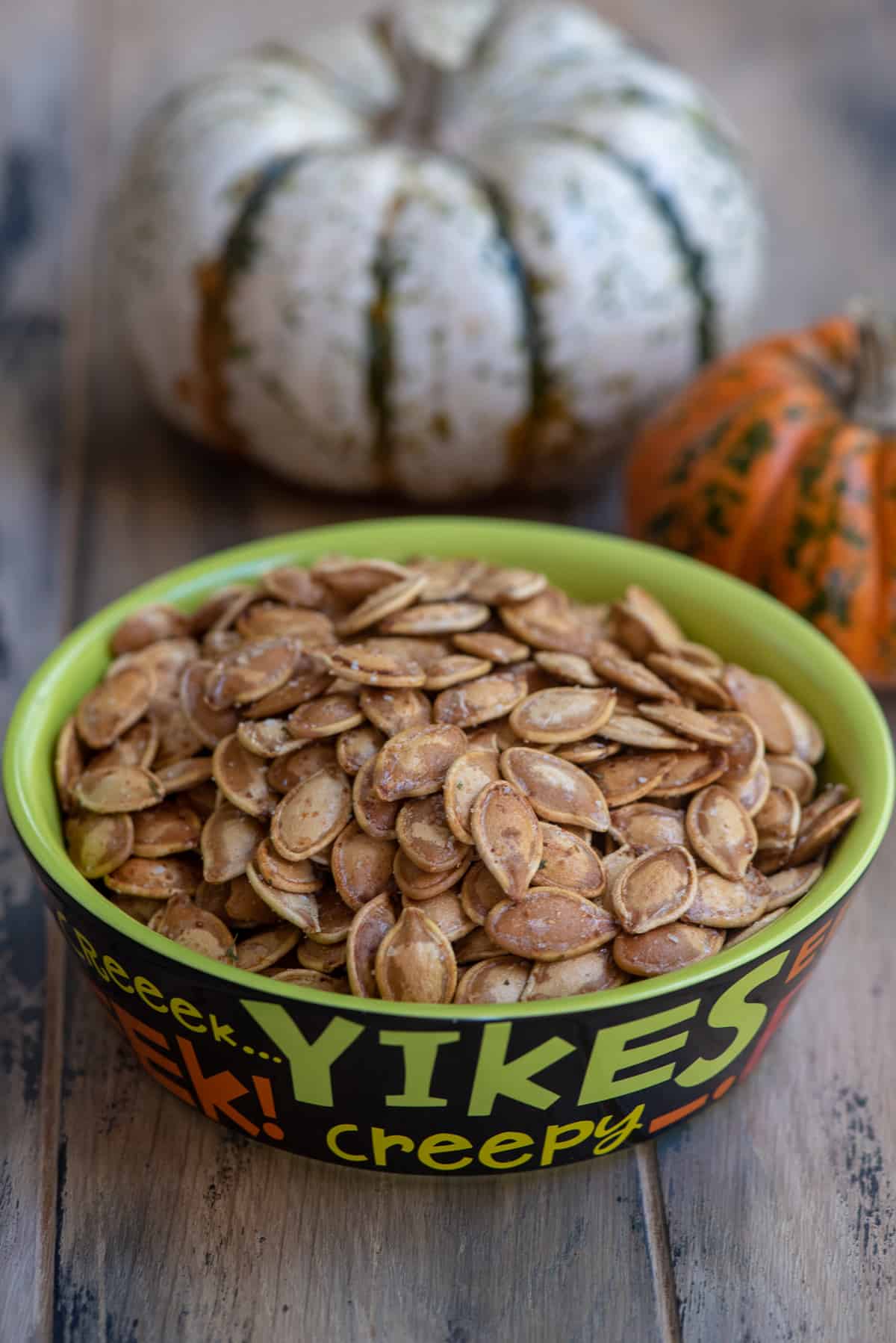 Roasted pumpkin seeds in a green bowl with pumpkins in the background.