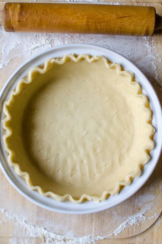 An unbaked pie crust in a white pie dish with a rolling pin behind it.