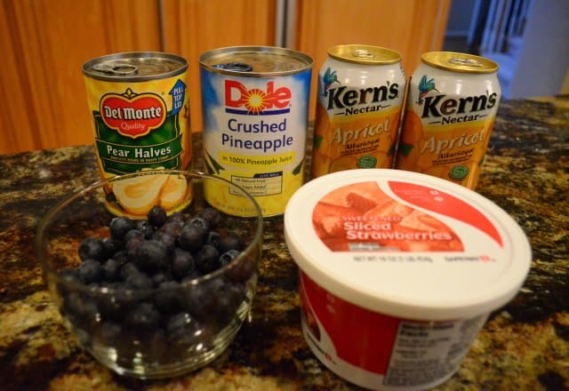 The ingredients on the kitchen counter include cans of apricot nectar (I use Kern's), frozen strawberries in syrup, a can of pears, a can of crushed pineapple, and fresh blueberries. 