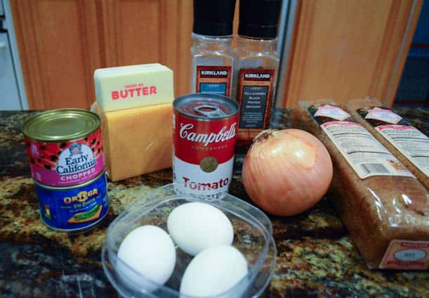 The ingredients - Sharp cheddar cheese, a stick of cold butter, 3  hard boiled eggs, a can of chopped olives, a can of diced green chiles, onion, a can of Campbell's Tomato Soup, and a little salt and black pepper - on a kitchen counter.
