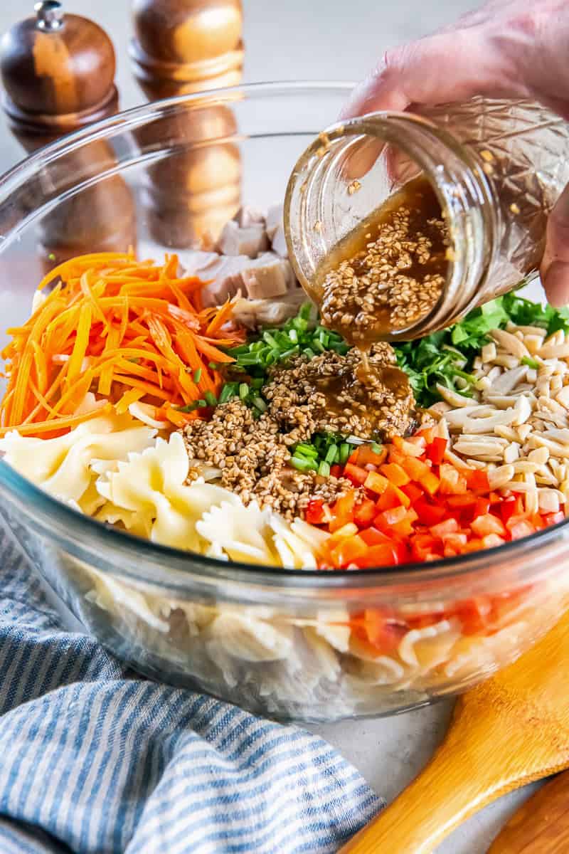 Pouring sesame dressing over the ingredients for Asian Pasta Salad in a large glass mixing bowl.