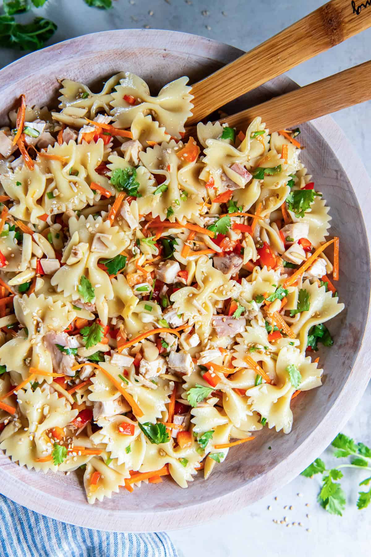 Asian Pasta Salad with bowtie pasta, chicken, carrots, red bell pepper, cilantro, and almonds in a wooden bowl with wooden salad servers.