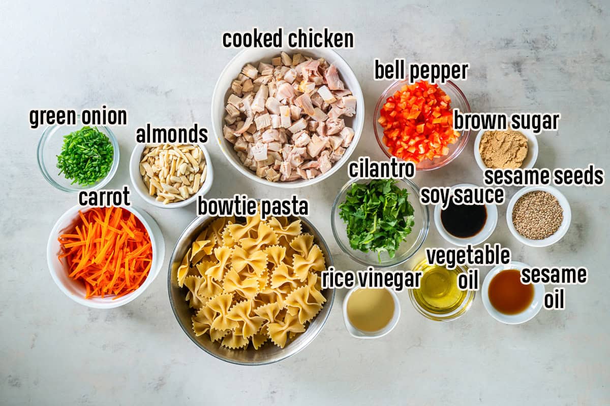 Ingredients for Asian Pasta Salad arranged in bowls, including bowtie pasta, cooked chicken, carrots, red bell pepper, green onion, cilantro, almonds, sesame seeds, soy sauce, rice vinegar, vegetable oil, sesame oil, and brown sugar.