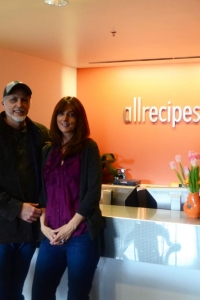 A man and a woman standing in a room in front of an allrecipes.com sign.