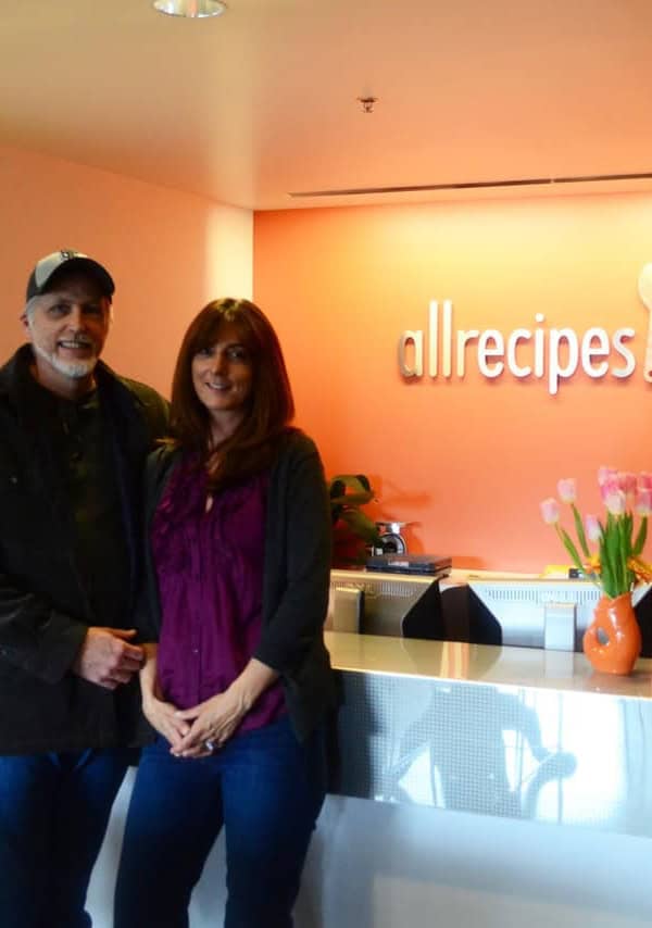 A man and a woman standing in a room in front of an allrecipes.com sign.