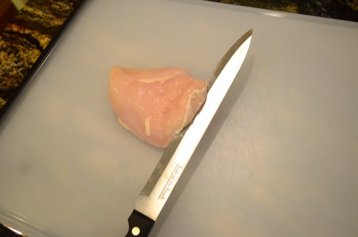 A raw chicken breast on a cutting board with a knife carefully cutting through the center of it.