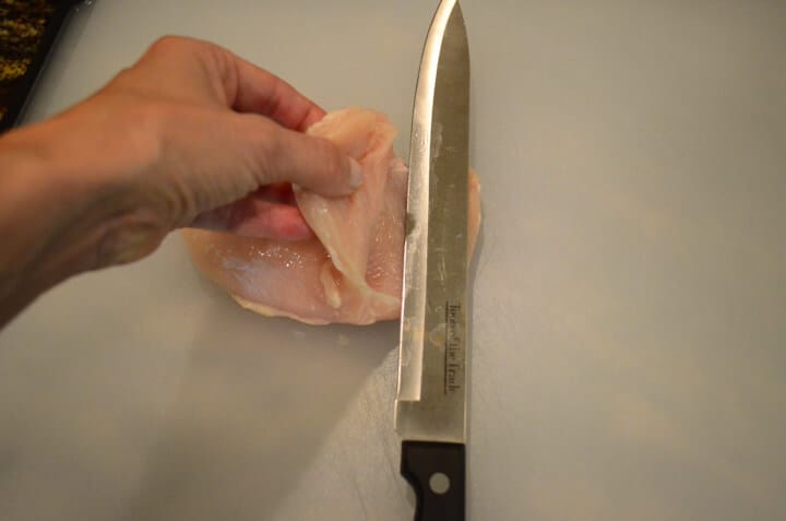 The raw chicken breast being cut through the middle with a hand holding the top half.