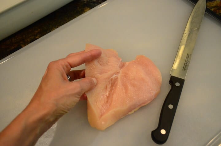 The raw chicken breast nearly cut in half with a hand folding it open.