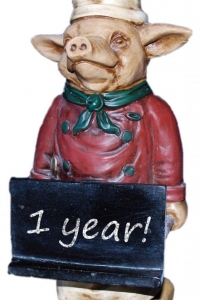 A ceramic pig dressed as a chef holding a chalkboard sign that says 1 year!