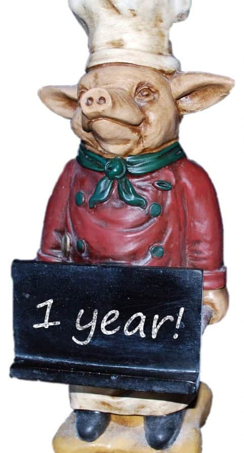 A ceramic pig dressed as a chef holding a chalkboard sign that says 1 year!