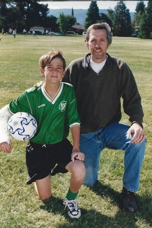 A man and his son holding a soccer ball kneeling on grass.