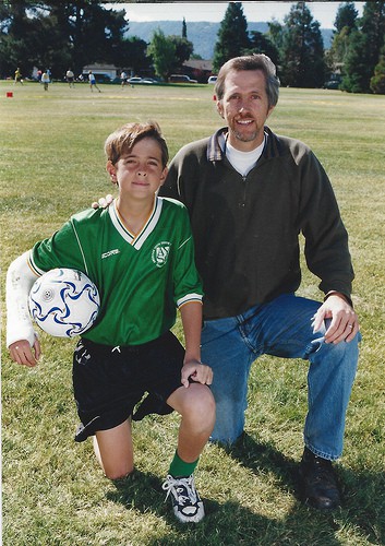 A man and his son holding a soccer ball kneeling on grass.