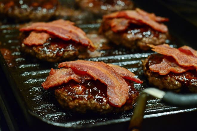 Stout & Sriracha Barbecue Bacon Burgers cooking on the grill with bacon on top.