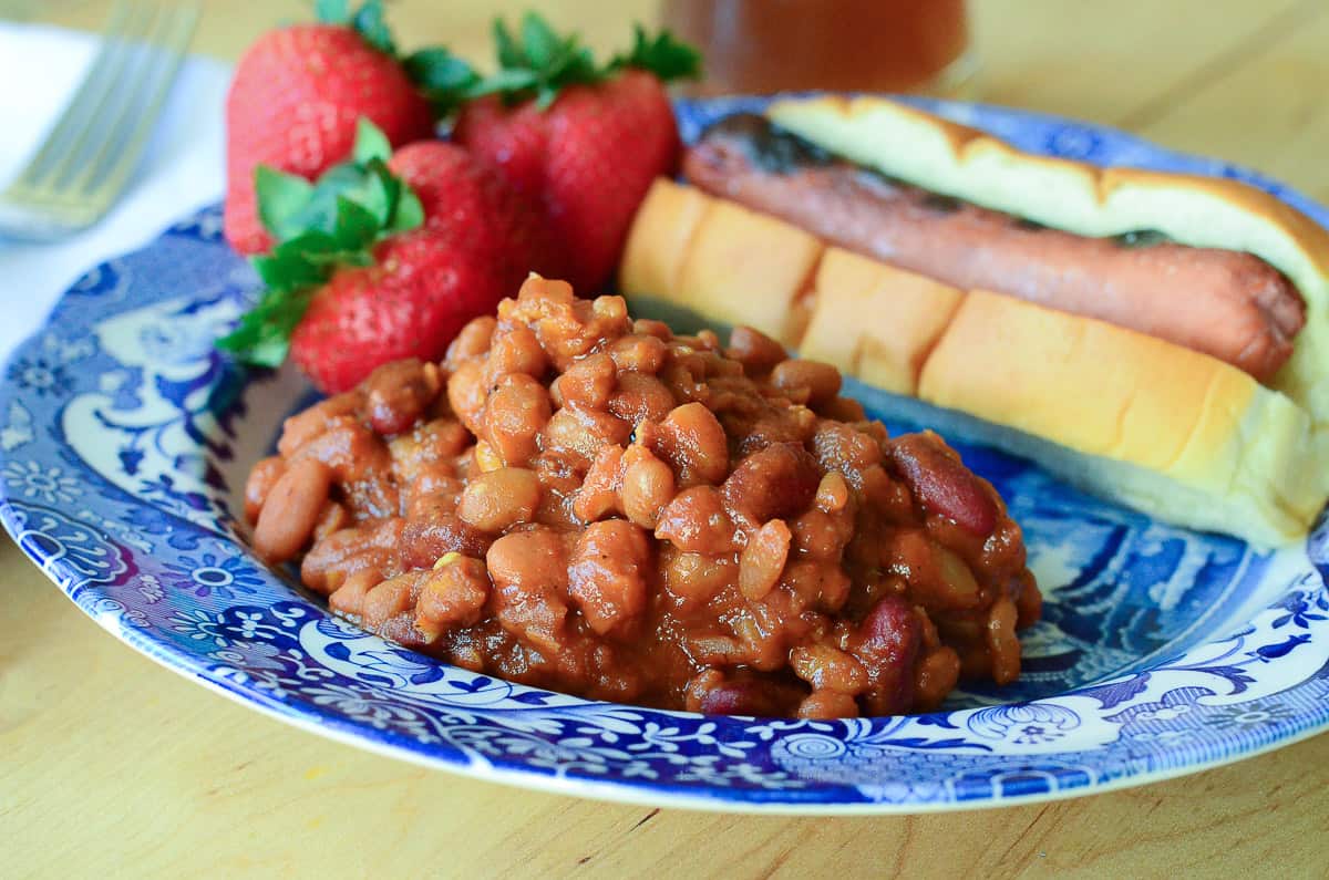 A serving of Spicy Baked Beans with a hot dog and strawberries on a blue plate.