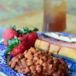 A serving of Spicy Baked Beans on a blue plate with a hot dog and strawberries.