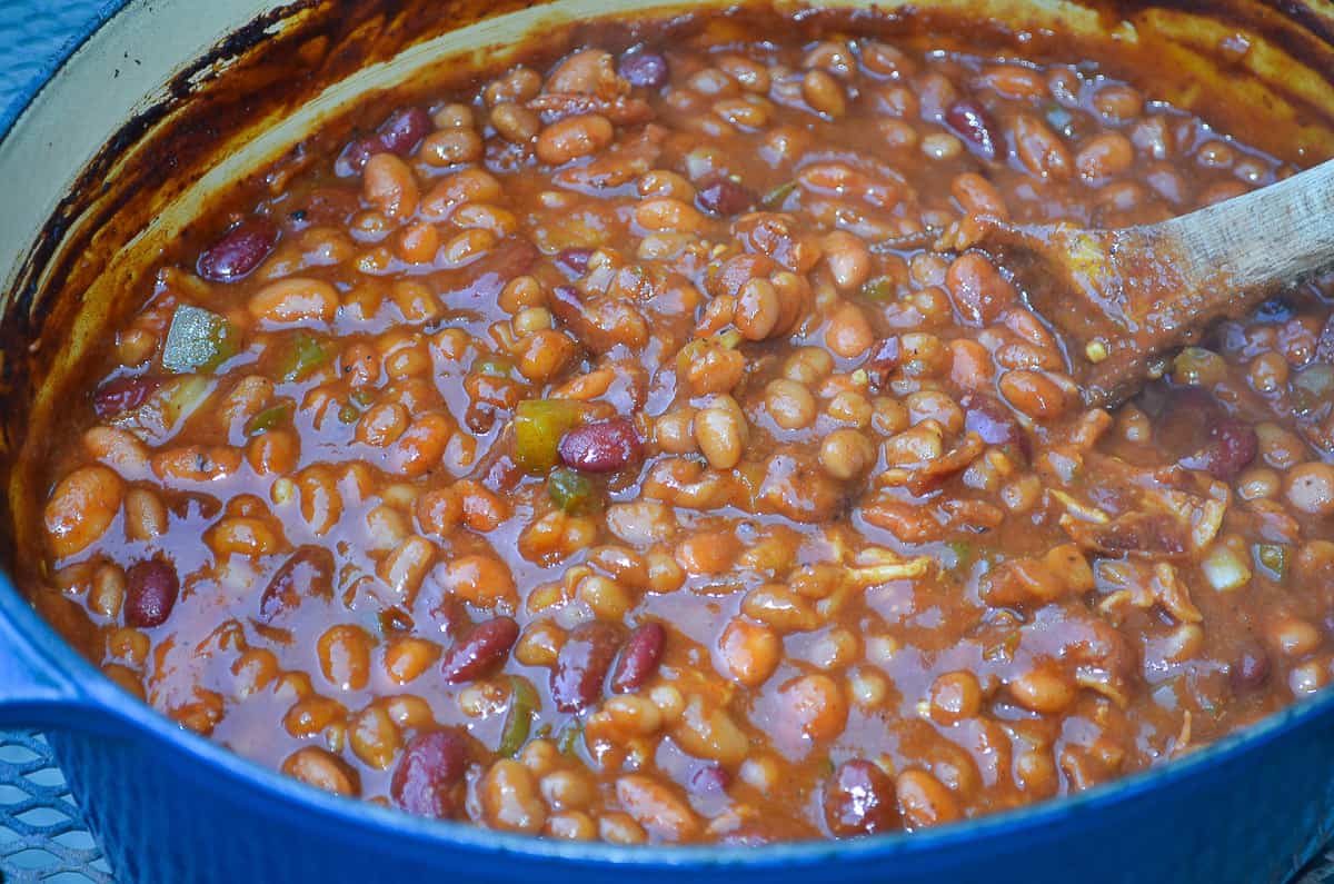 The finished Spicy Baked Beans in a blue Dutch oven with a wooden spoon.