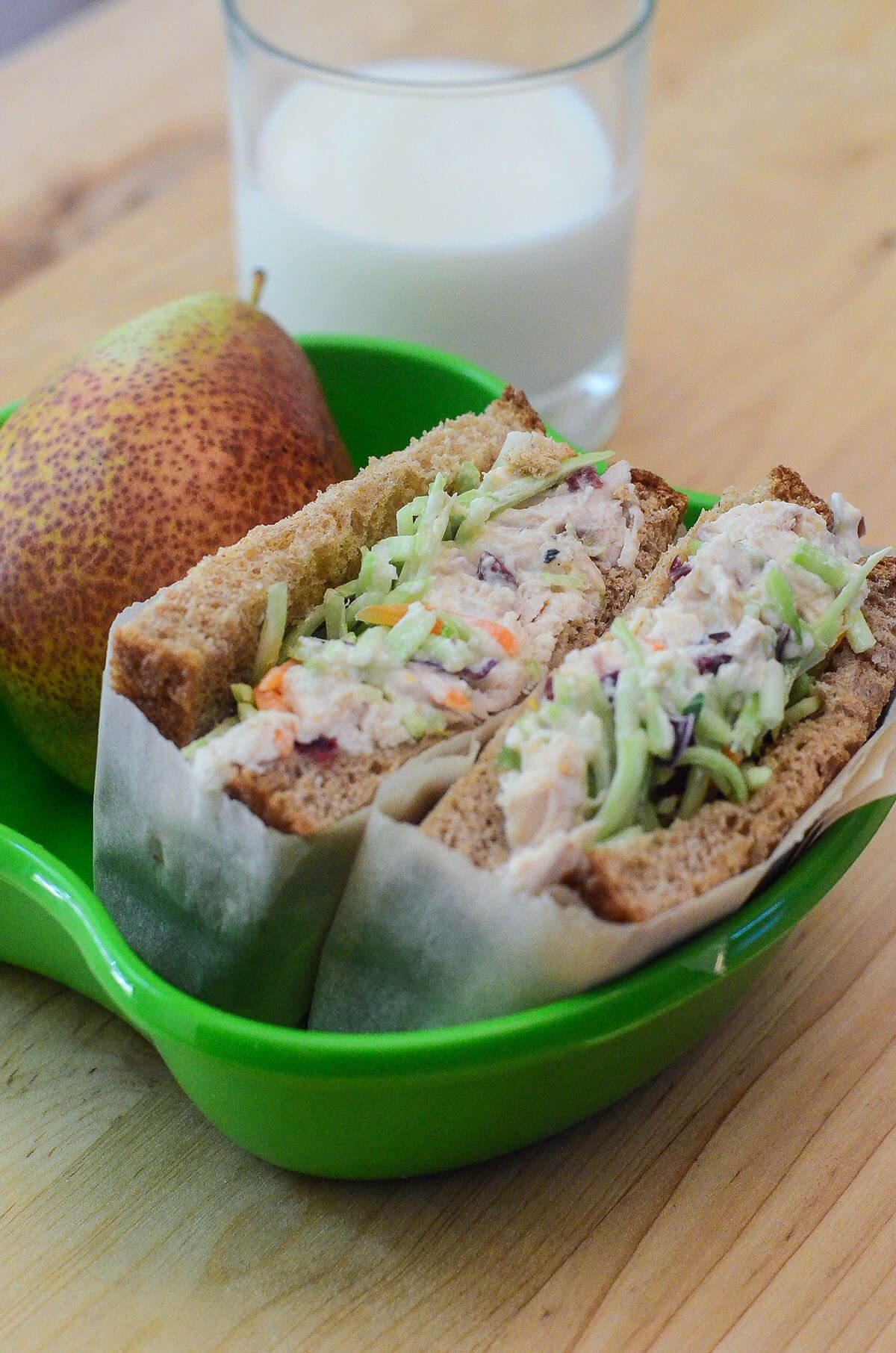 A sandwich and a pear in a green bowl with a glass of milk behind it.