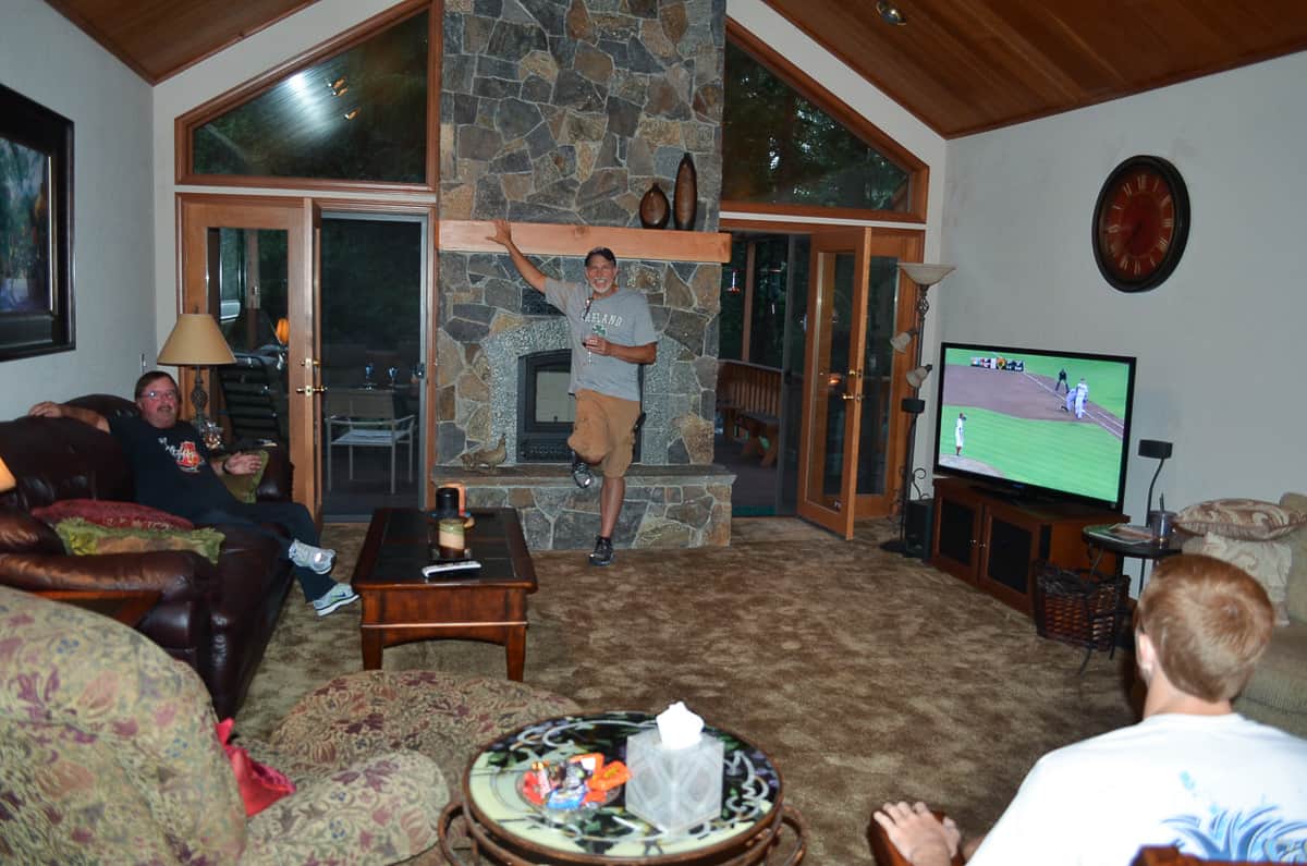 A man leans against a fireplace wall while two other men sit in a living room.