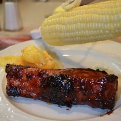 A knife smears butter on corn on the cob hovering over a plate with a Grilled Country Style Rib.