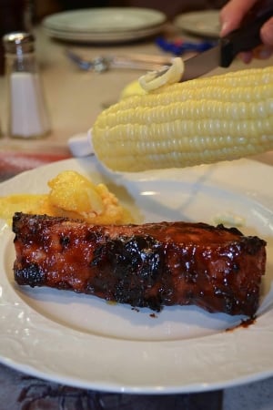 A knife smears butter on corn on the cob hovering over a plate with a Grilled Country Style Rib.