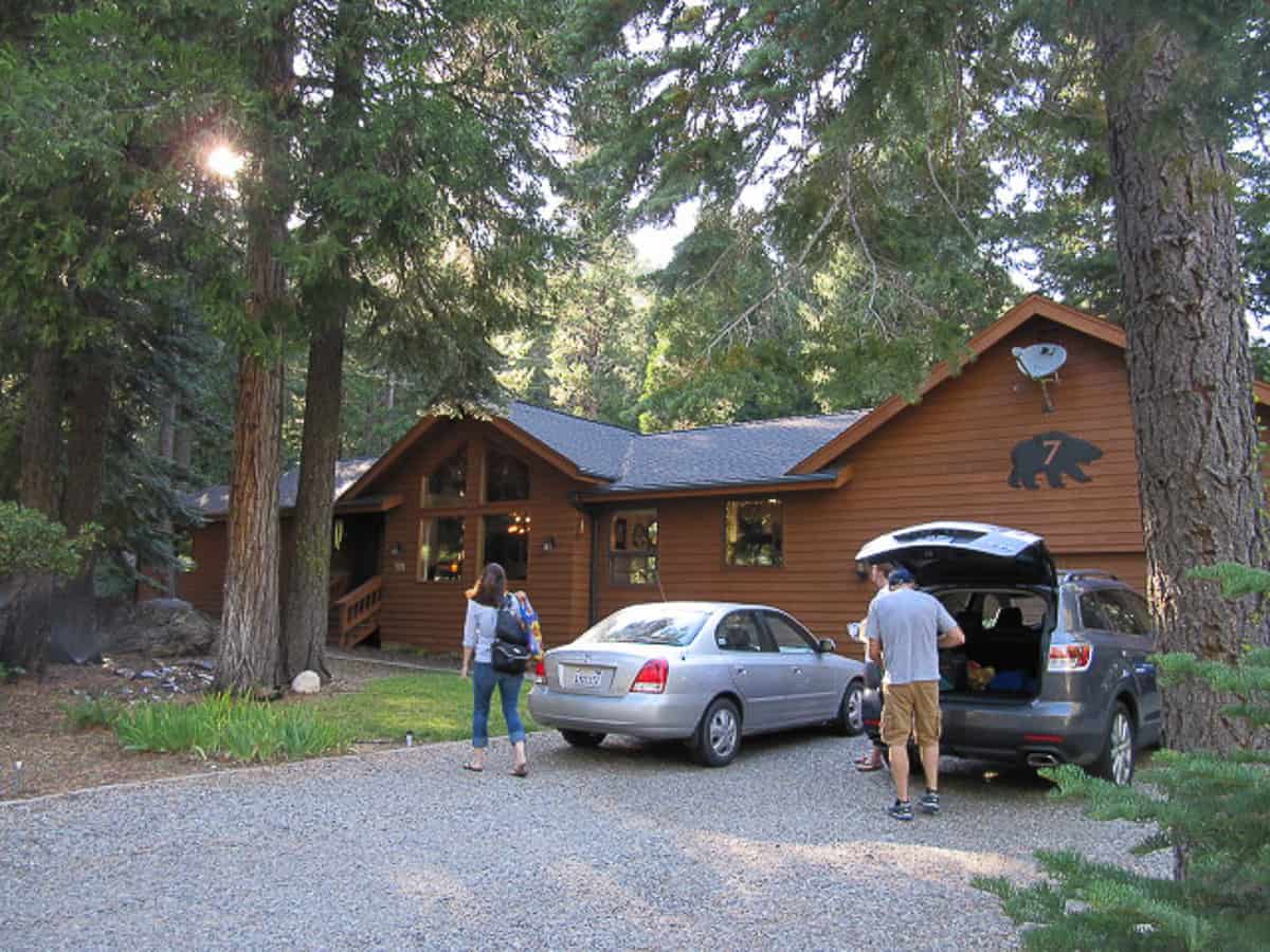 A man unloading the trunk of a car in a driveway and a woman walking towards a cabin.