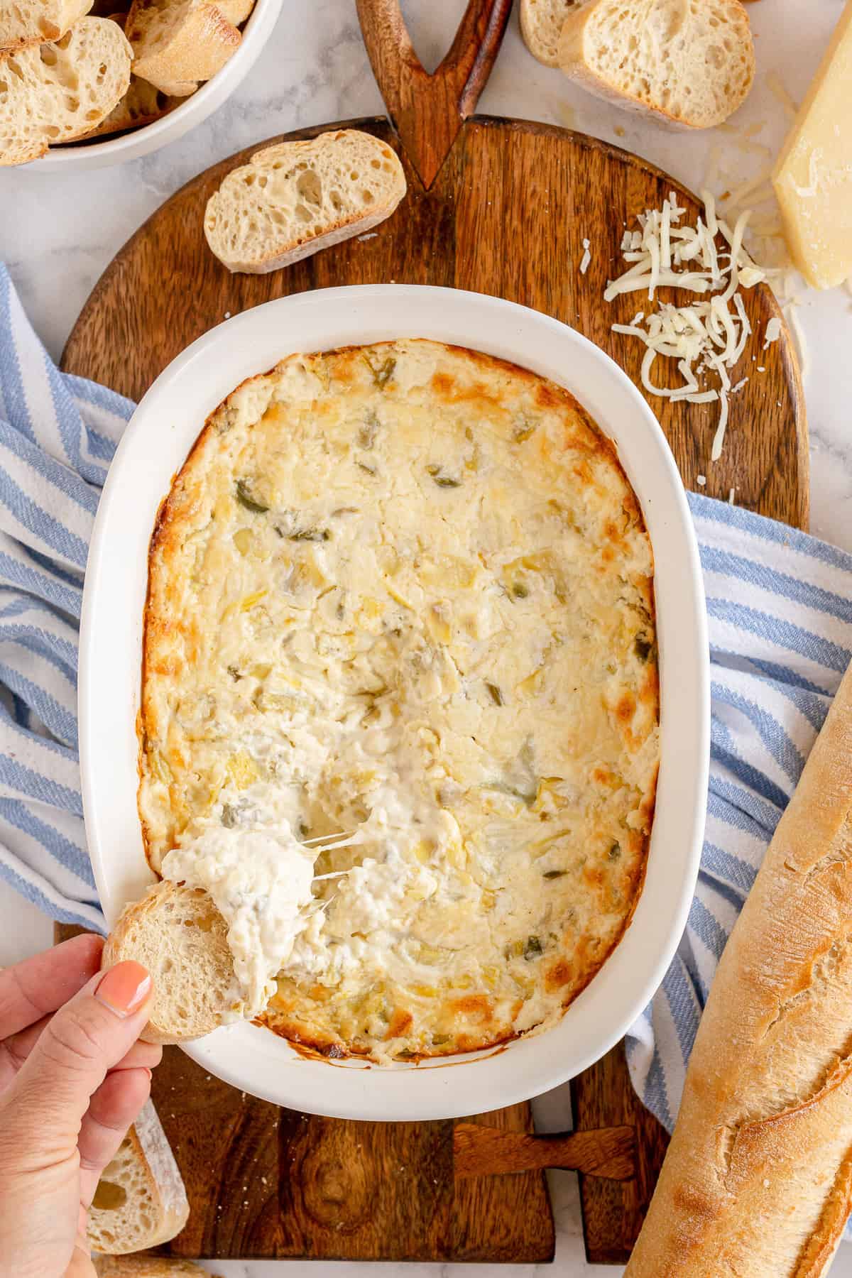 An over the top shot of a hand dipping a baguette slice into Artichoke Dip.