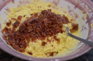 Crumbled bacon is added to the deviled egg mixture in a small mixing bowl.