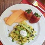 A plate of scrambled eggs with black beans, avocado and sour cream next to fruit.
