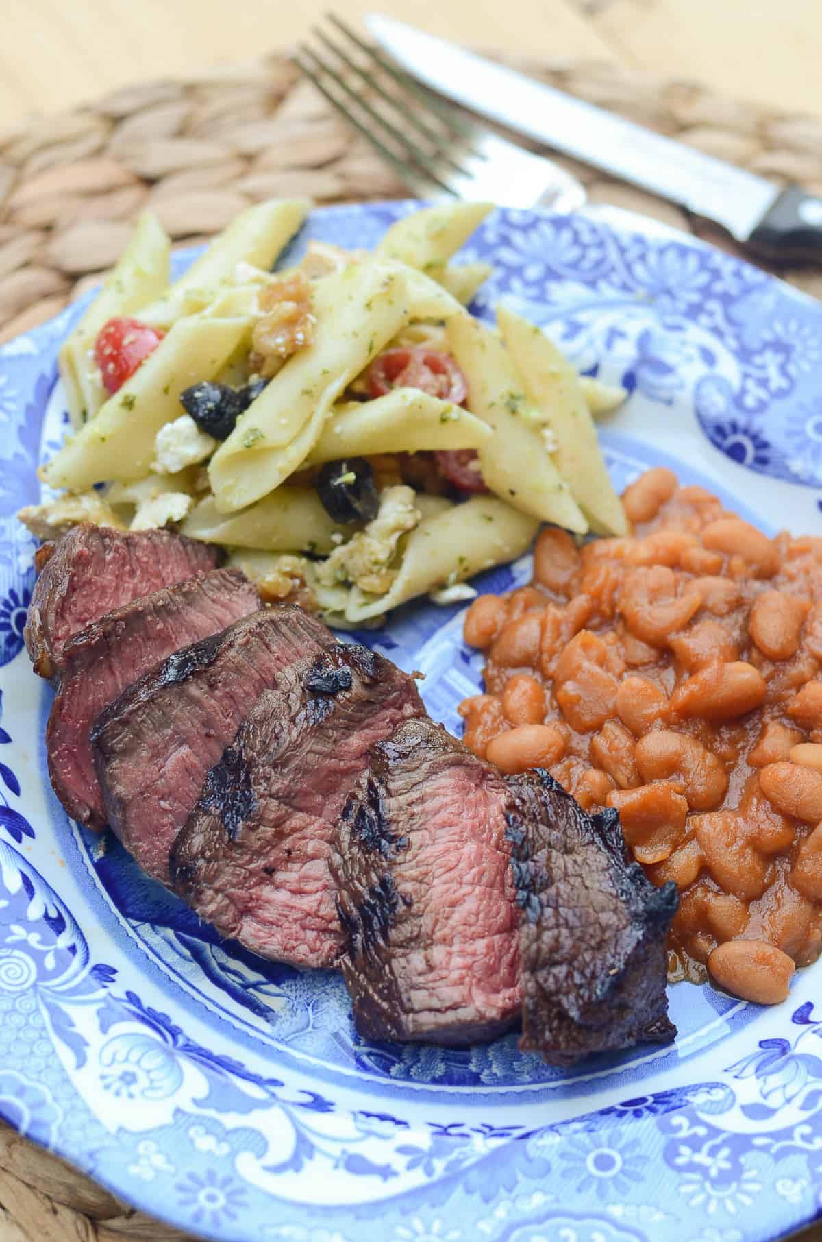 Sliced grilled steak on a blue plate with pasta salad and beans.