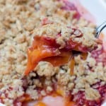 A spoon scoops up some Peach Strawberry Crisp from a white baking dish.
