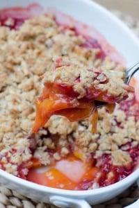 A spoon scoops up some Peach Strawberry Crisp from a white baking dish.