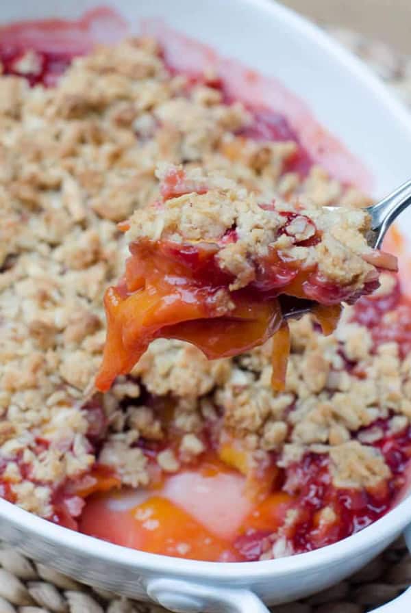 A spoon scoops up some Peach Strawberry Crisp from a white baking dish.