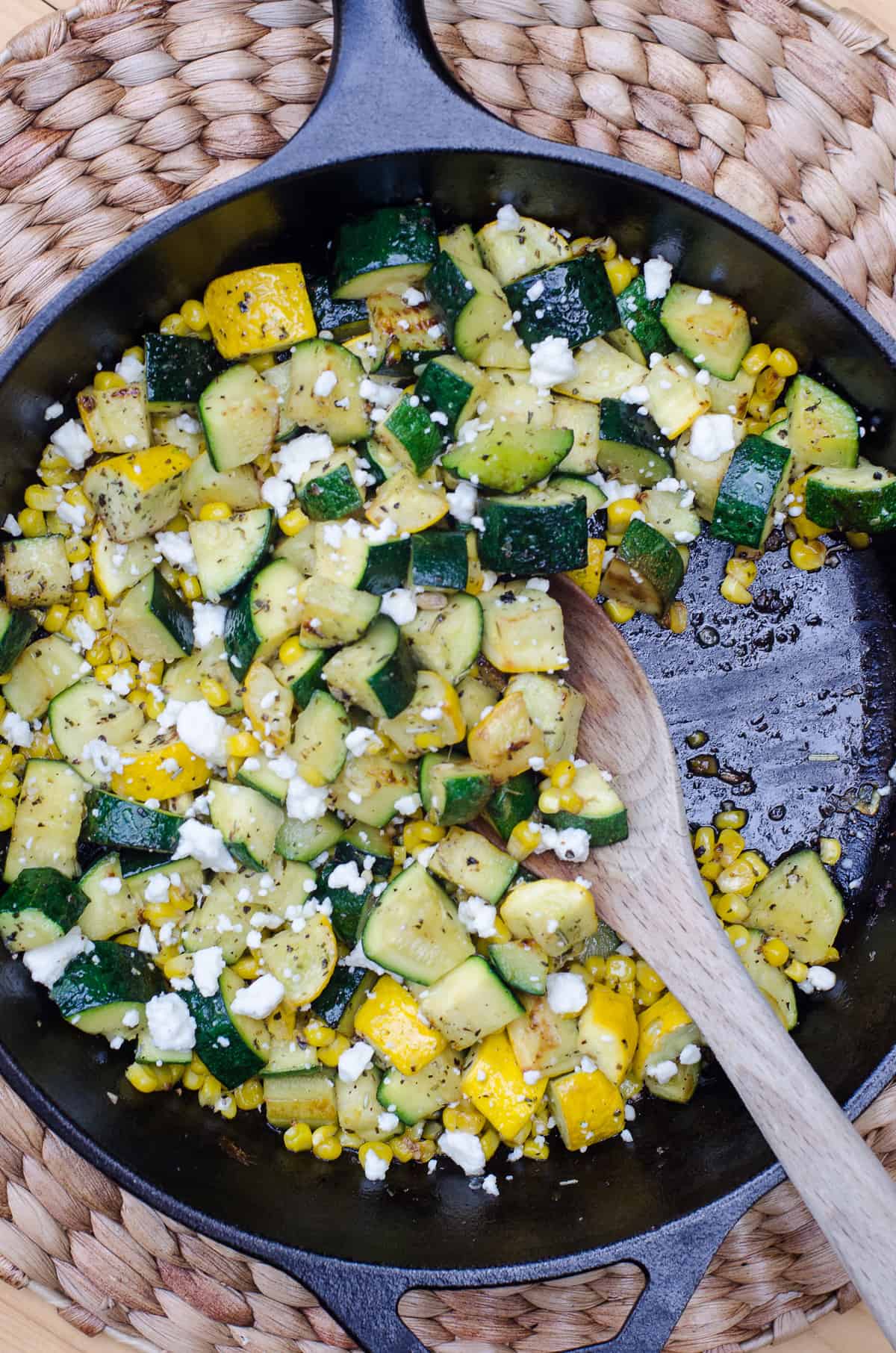 A cast iron skillet filled with cooked zucchini and corn and a wooden spoon shot from over the top.