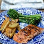 A piece of teriyaki salmon on a blue and white plate with french fries and broccolini.