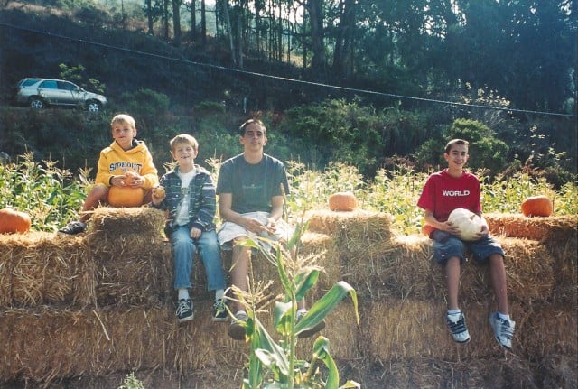 Four young boys sitting on bales of hay at a pumpkin patch.