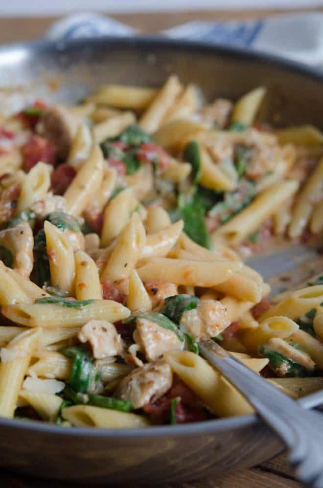 A closeup of penne with chicken and spinach and tomatoes in a skillet.