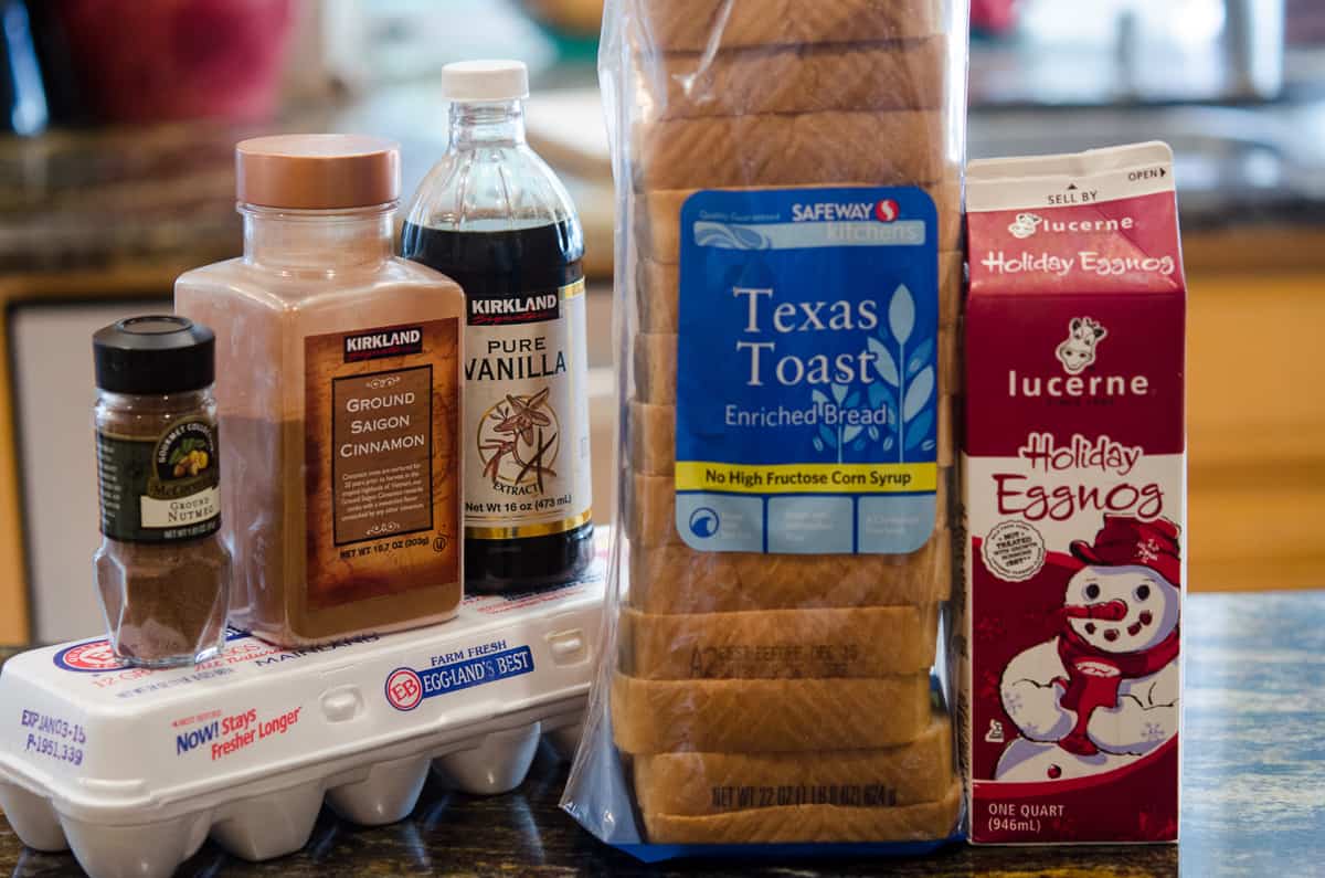 A carton of eggnog, sandwich bread, eggs, vanilla, cinnamon, and nutmeg on a kitchen counter.