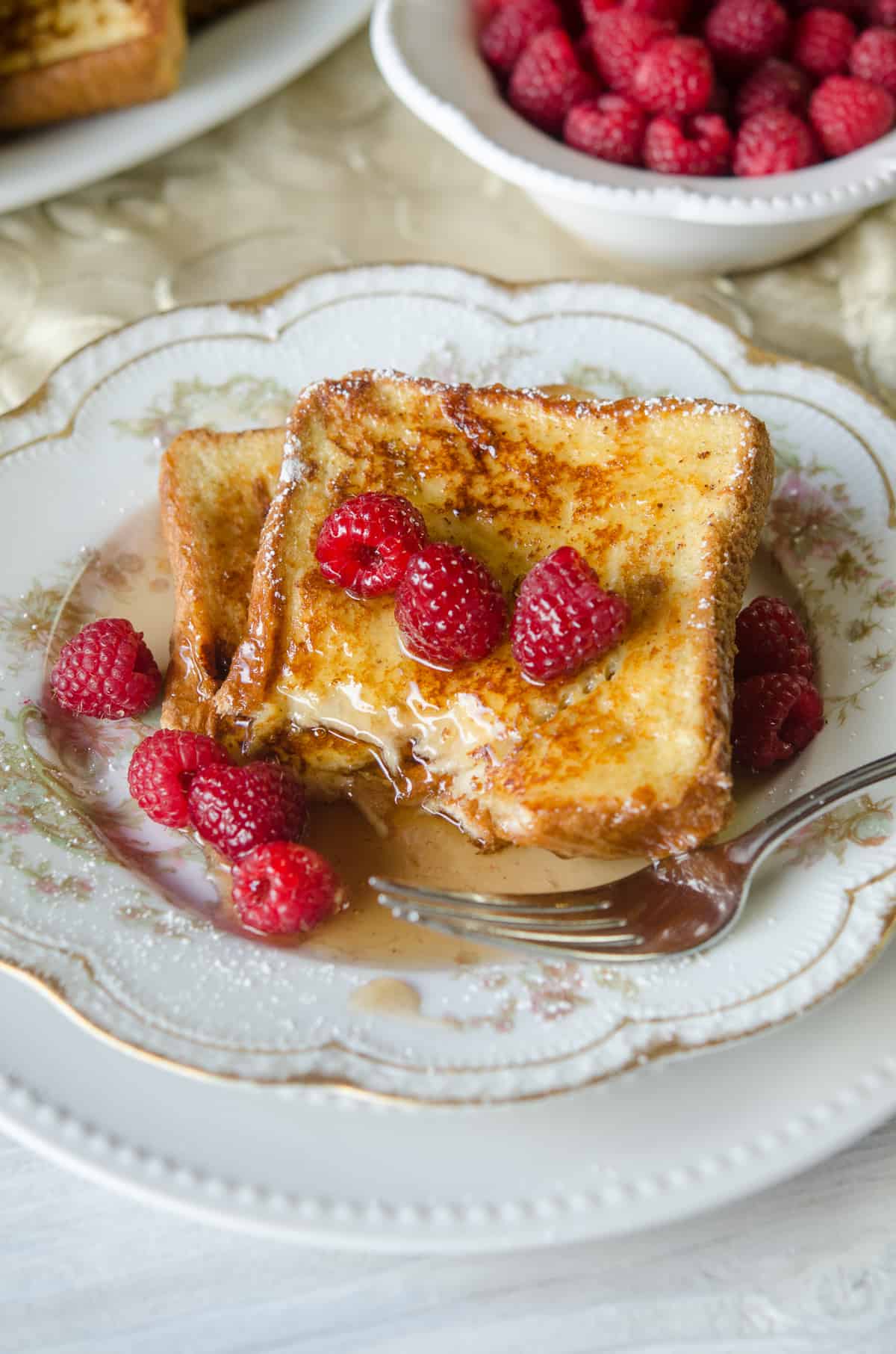 Eggnog French Toast topped with maple syrup with a bite missing on a china plate with raspberries and a fork.