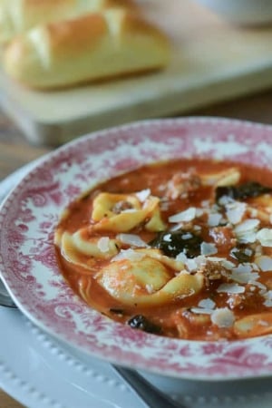 A red and white bowl filled with sausage and tortellini soup with kale.