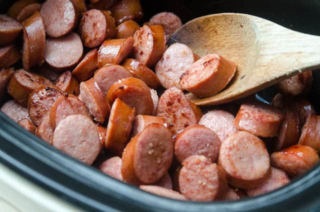 A wooden spoon stirs sausage in a slow cooker.