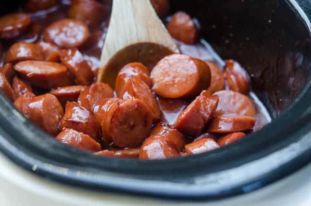 A wooden spoon stirs sausage into sauce in a crock pot.
