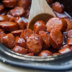 A wooden spoon stirs sausage in a slow cooker.