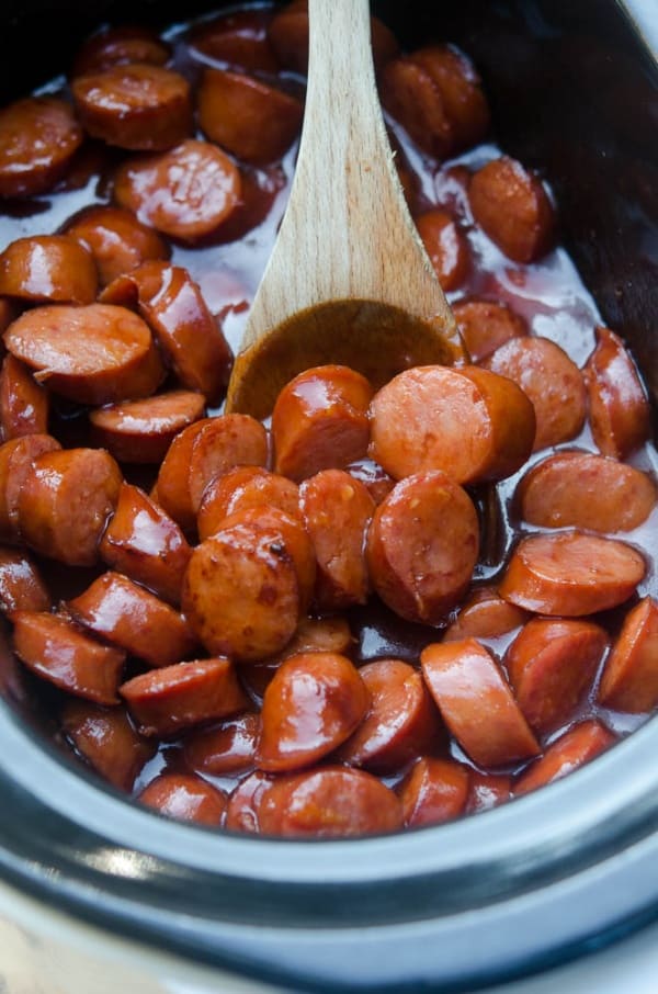 A wooden spoon stirs cocktail sausage in a bbq sauce in a slow cooker.