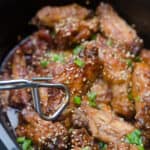 Metal tongs holding a Asian chicken wing over a slow cooker.