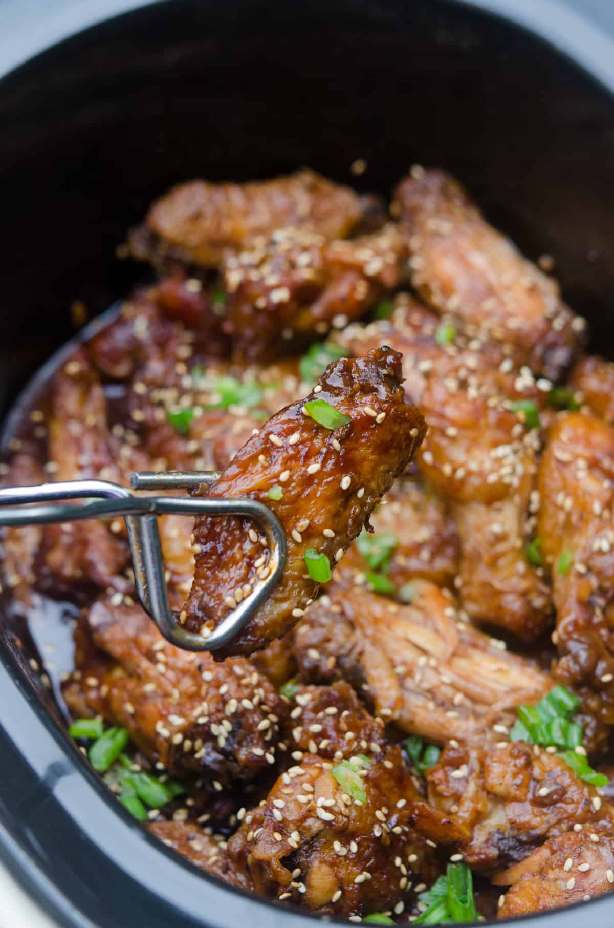 Metal tongs holding a Asian chicken wing over a slow cooker.