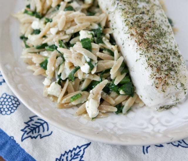 Orzo with spinach and feta with a piece of fish on a white plate.