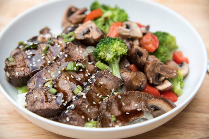 Slices of Teriyaki Steak in a white bowl with rice and veggies.