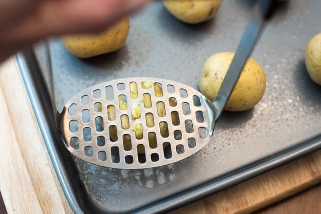 A metal potato masher smashes one of the potatoes on the baking sheet.