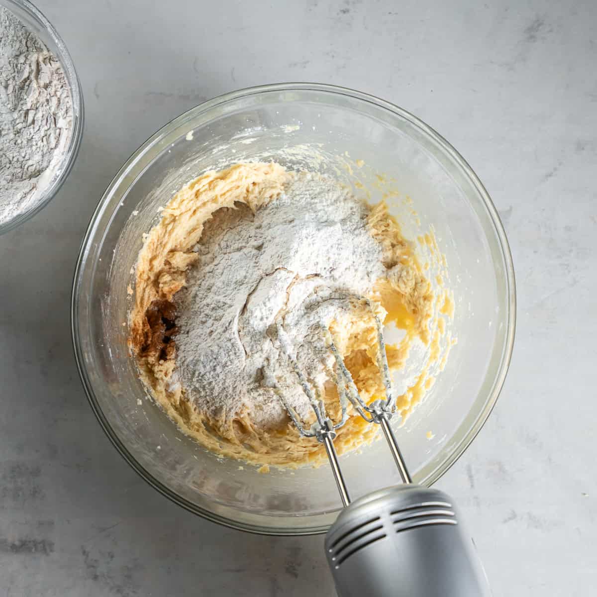 Flour mixture added to the wet ingredients in a glass bowl with an electric hand mixer.